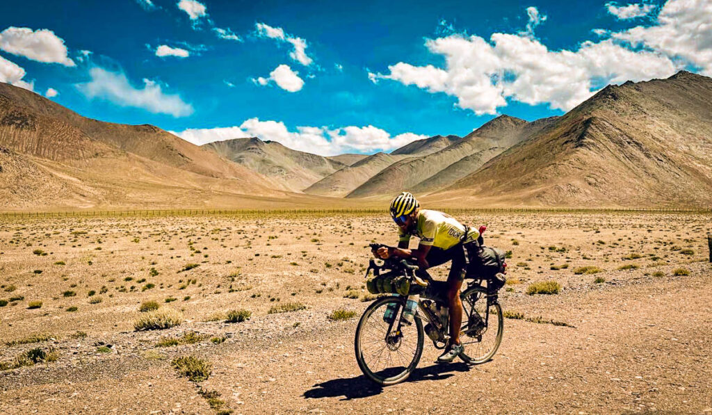Cyclist riding a fully loaded touring bike across a wide high-altitude valley on the Pamir Highway