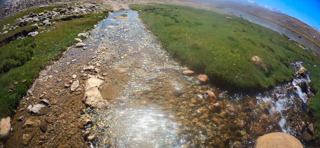 Shallow rocky stream crossing a green meadow on the Zorkul detour, Pamir Highway